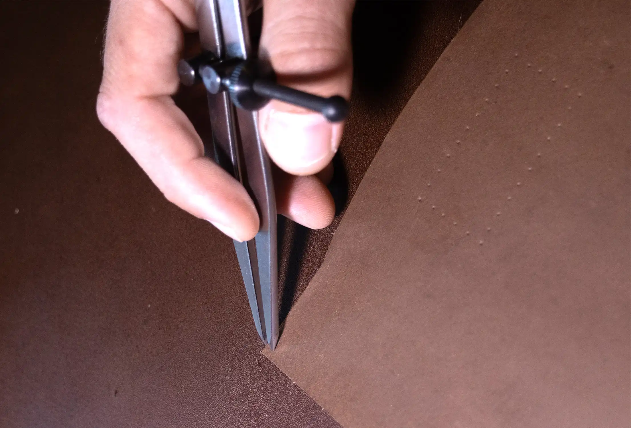 A leather working using a compass to mark the edge of a piece of brown leather.