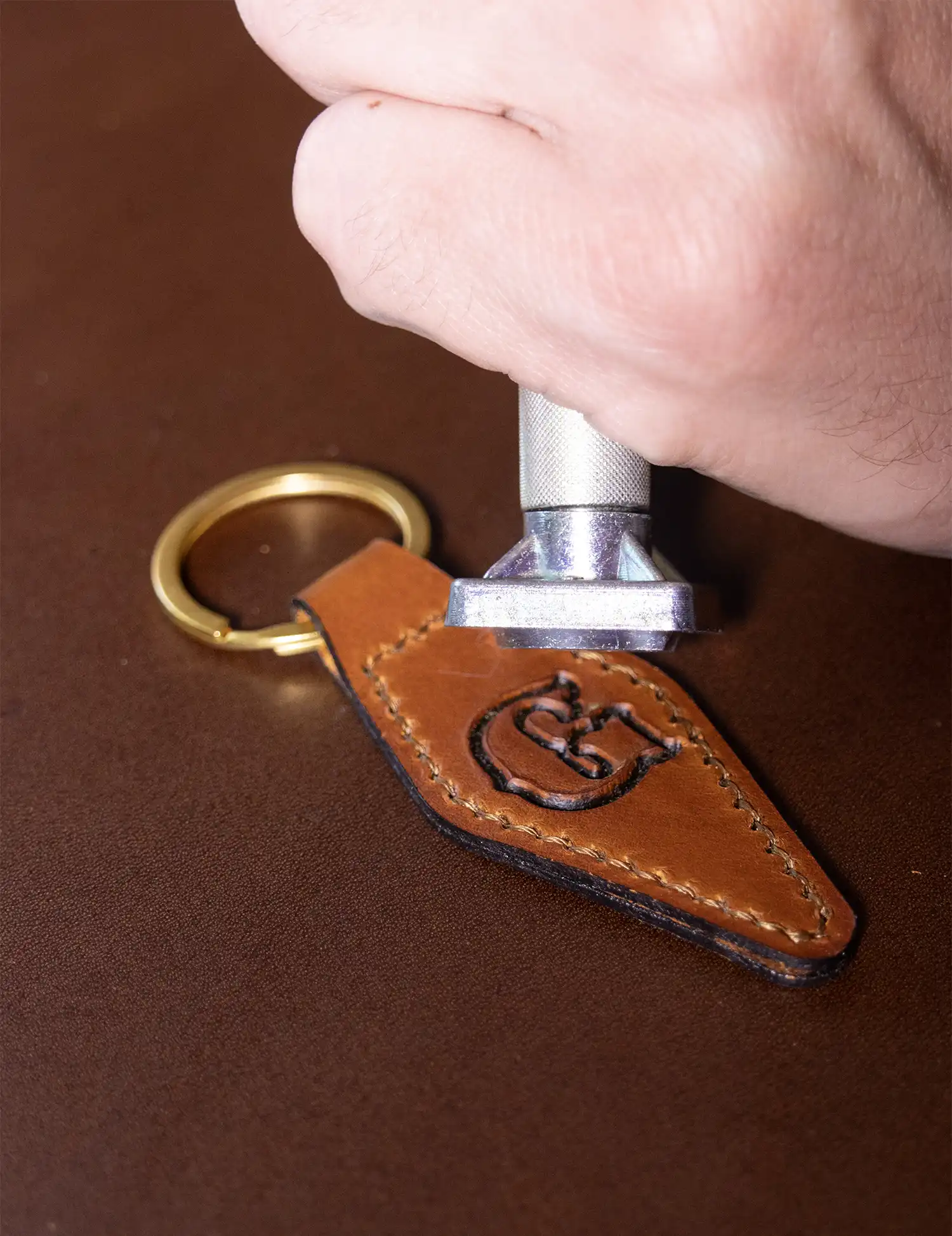 Leather worker stamping the letter G into a brown leather keychain that has a brozne keyring.
