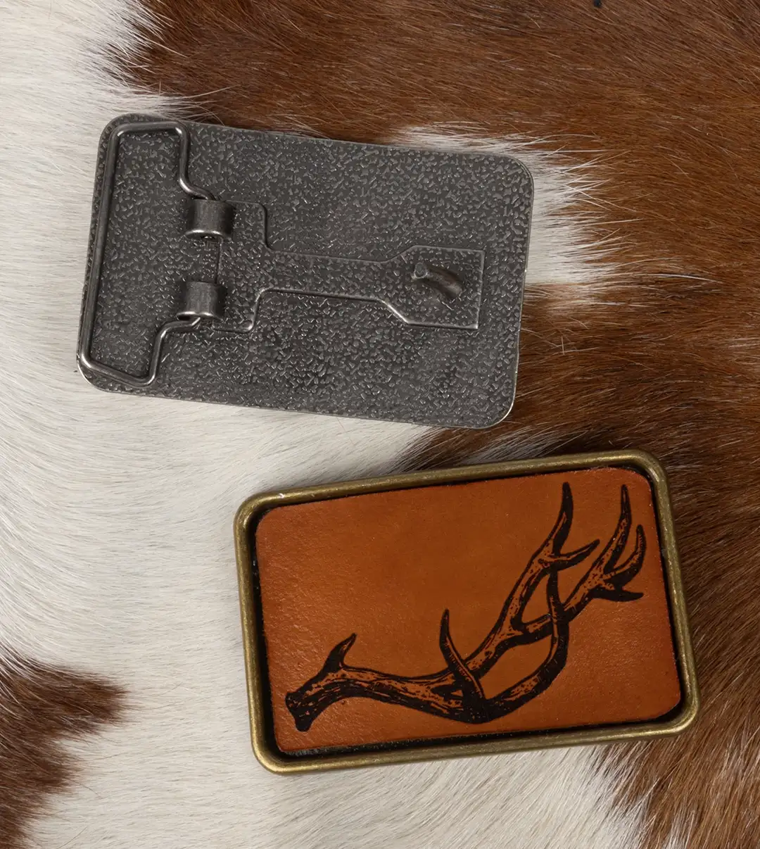 A bronze belt buckle featuring a laser engraved leather inlay of an antler displayed on a brown and white pelt.