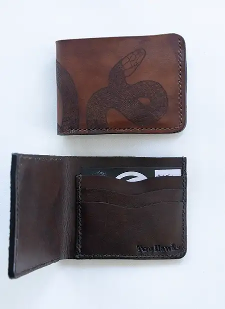 A brown leather bi-fold wallet with a hand-stamped blacksnake design on the front cover displayed on a neutral surface.