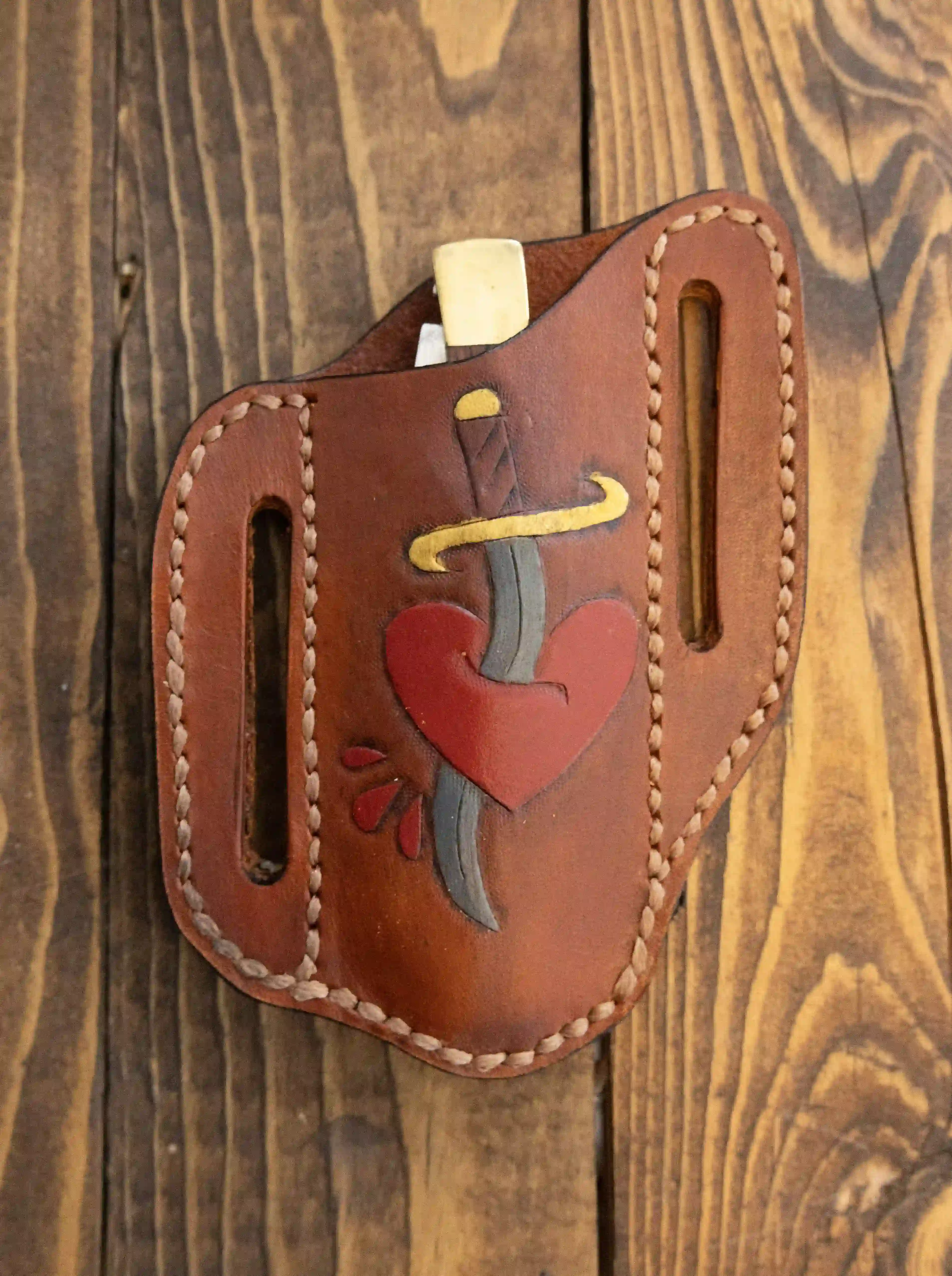 A slim pancake-style leather knife sheath with a hand-embossed dagger piercing a heart on the front face displayed on a wooden surface.