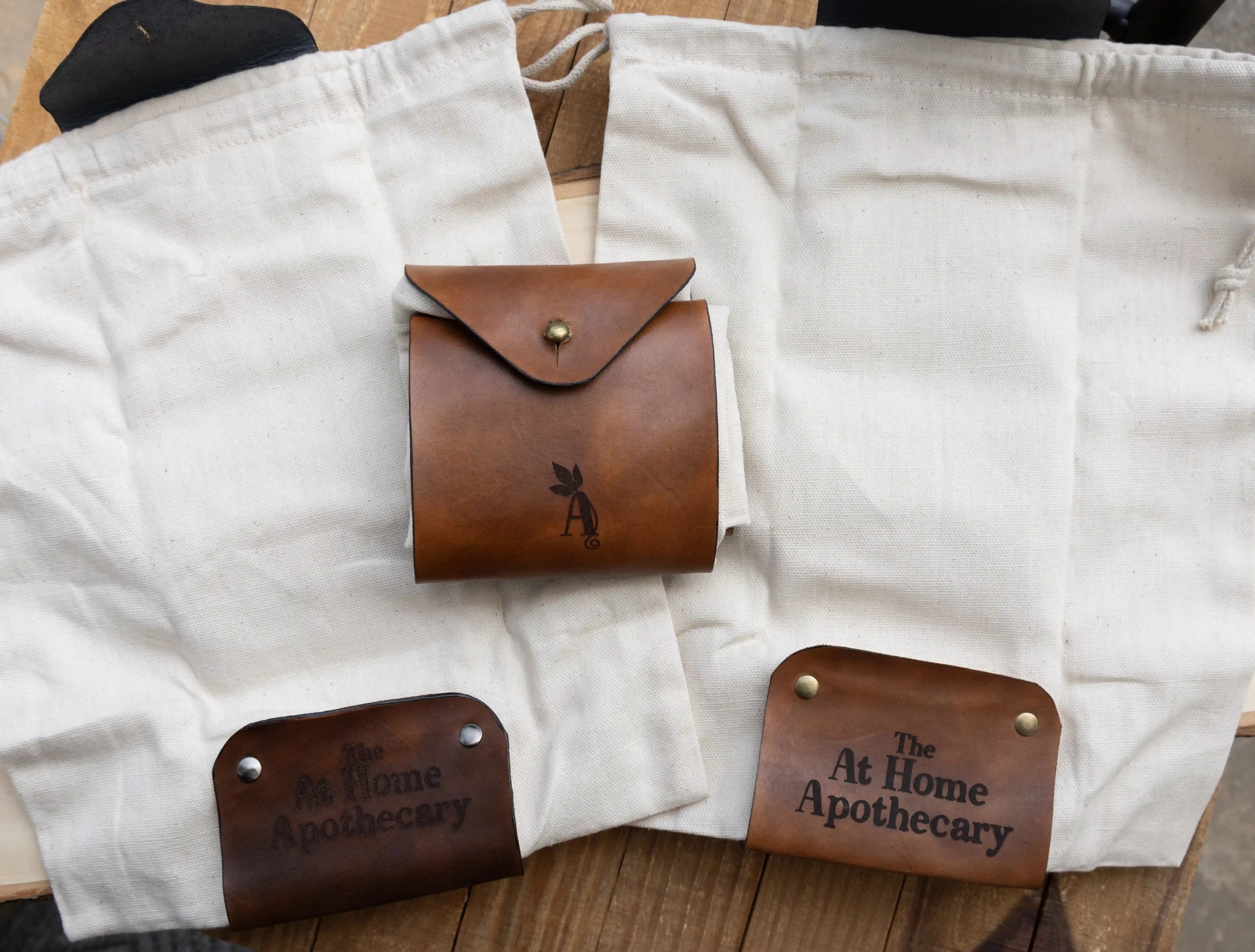 Two open and one closed white canvas foraging pouches featuring a hand-embossed leather cuff with bronze hardware displayed on a live edge wooden plank.