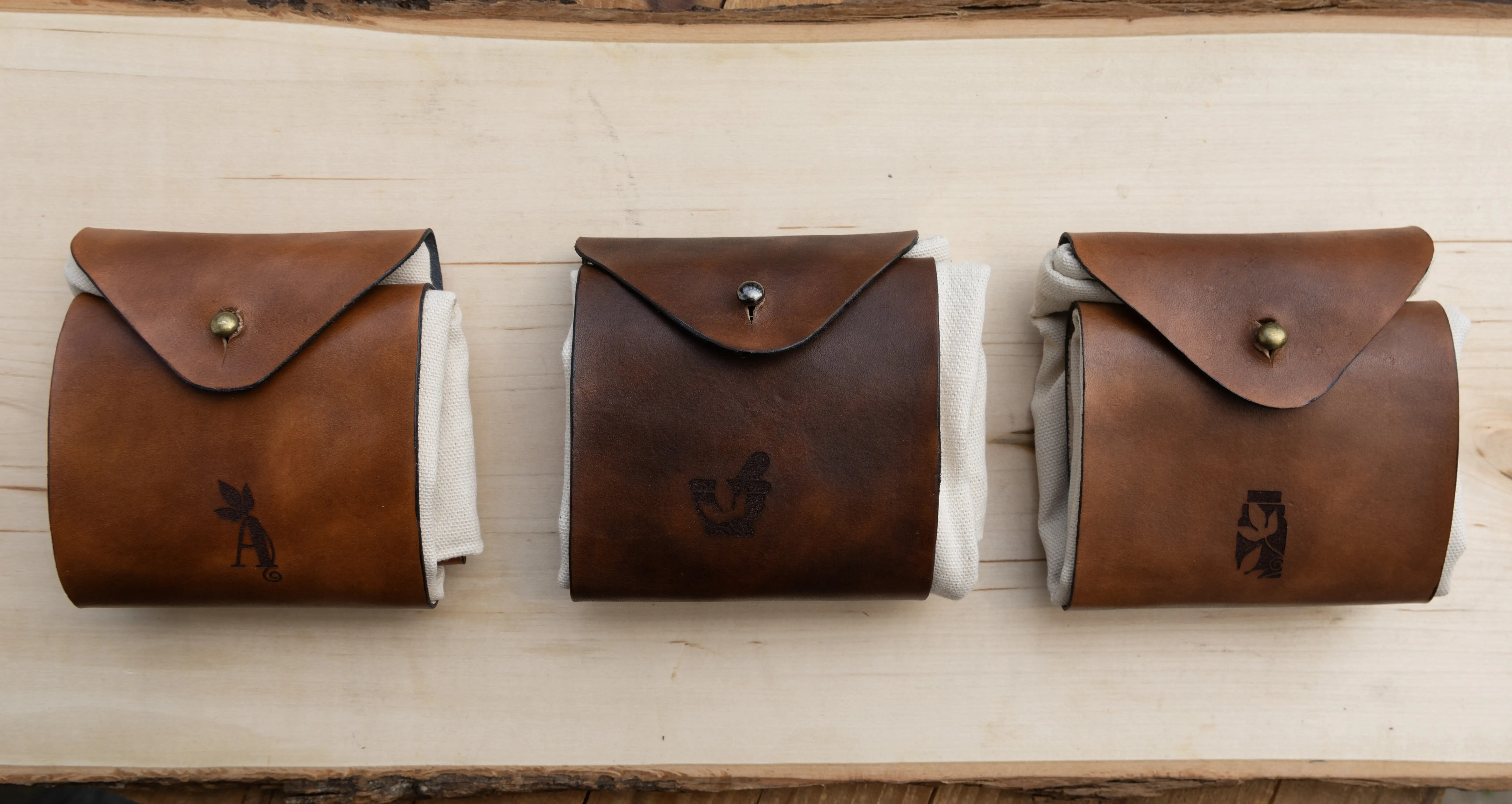 Three closed foraging pouches displayed together — two with bronze hardware and one with silver hardware displayed on a wooden table.