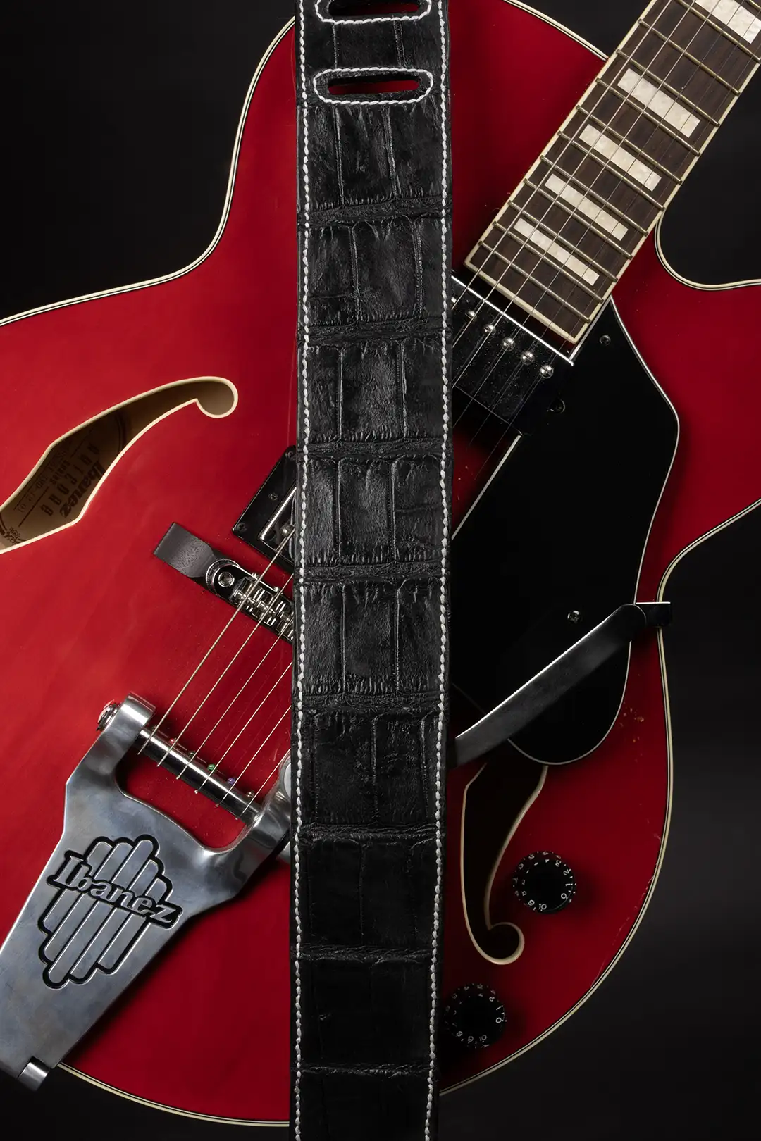 Black full-grain leather guitar strap with hand-embossed gator texture and white contrast stitching, shown on a red electric guitar.