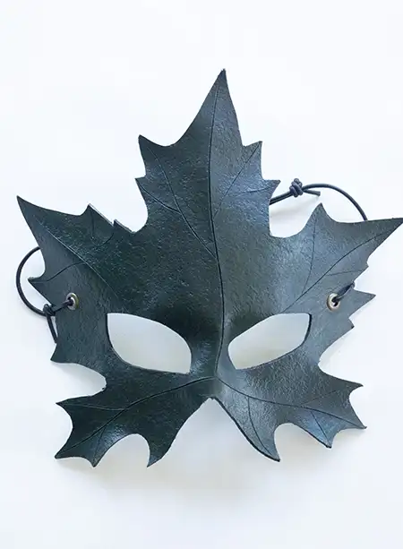 A green leather mask shaped like a maple leaf with an adjustable black leather strap displayed against a neutral background