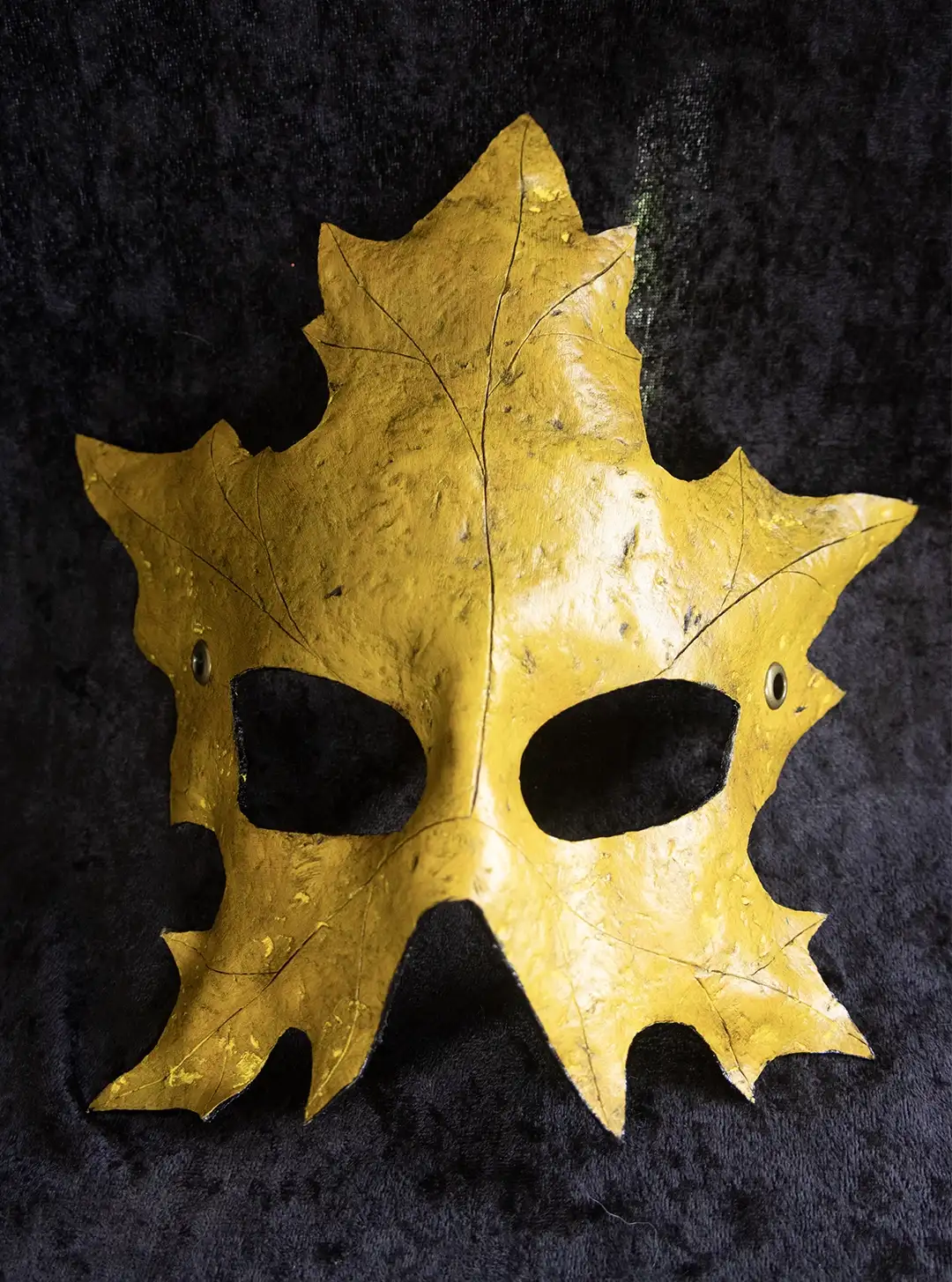A yellow leather mask shaped like a maple leaf with an adjustable black leather strap displayed against a dark background.