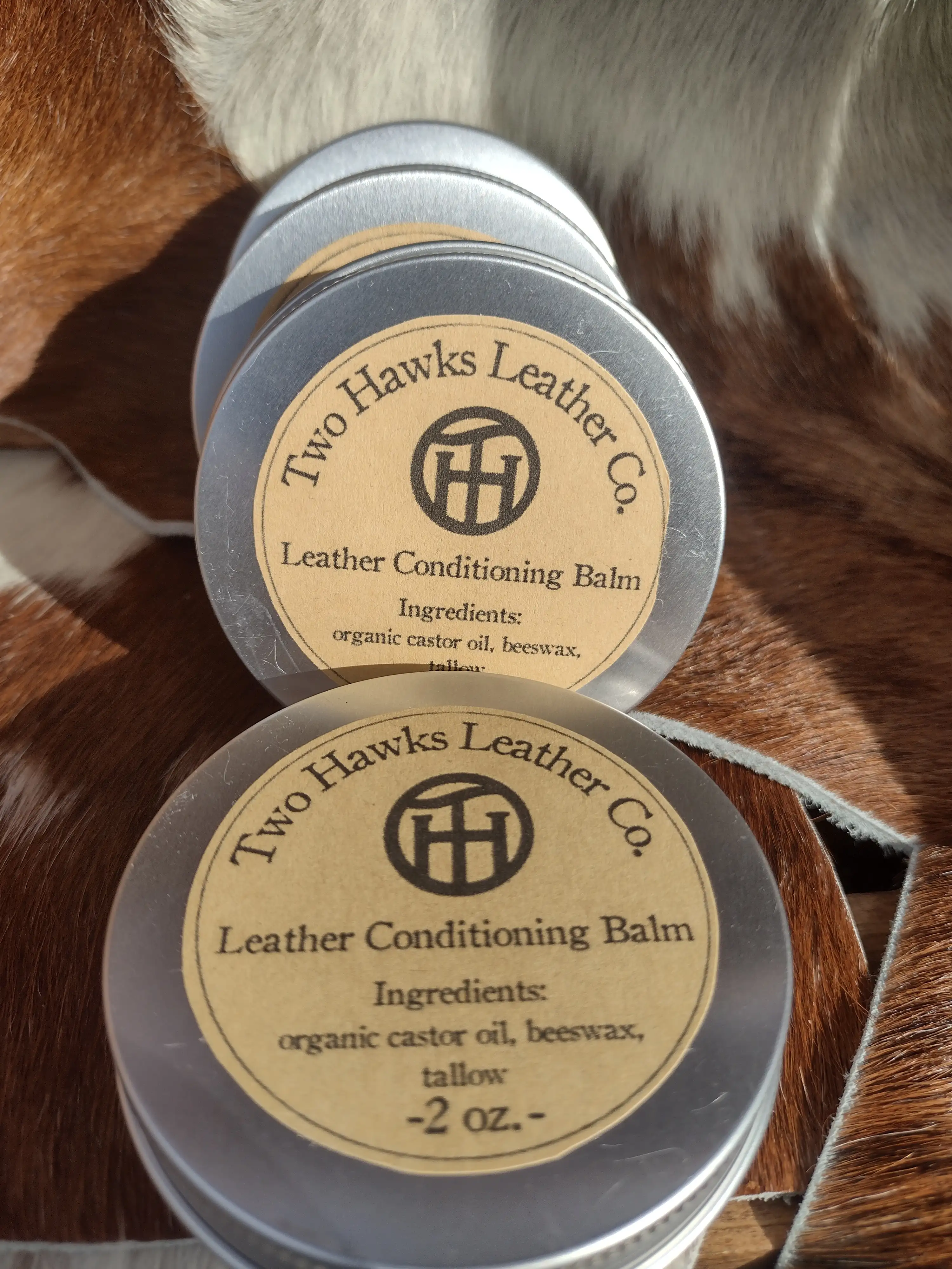 Multiple 2-ounce tins of Two Hawks Leather balm displayed on brown and white pelts.