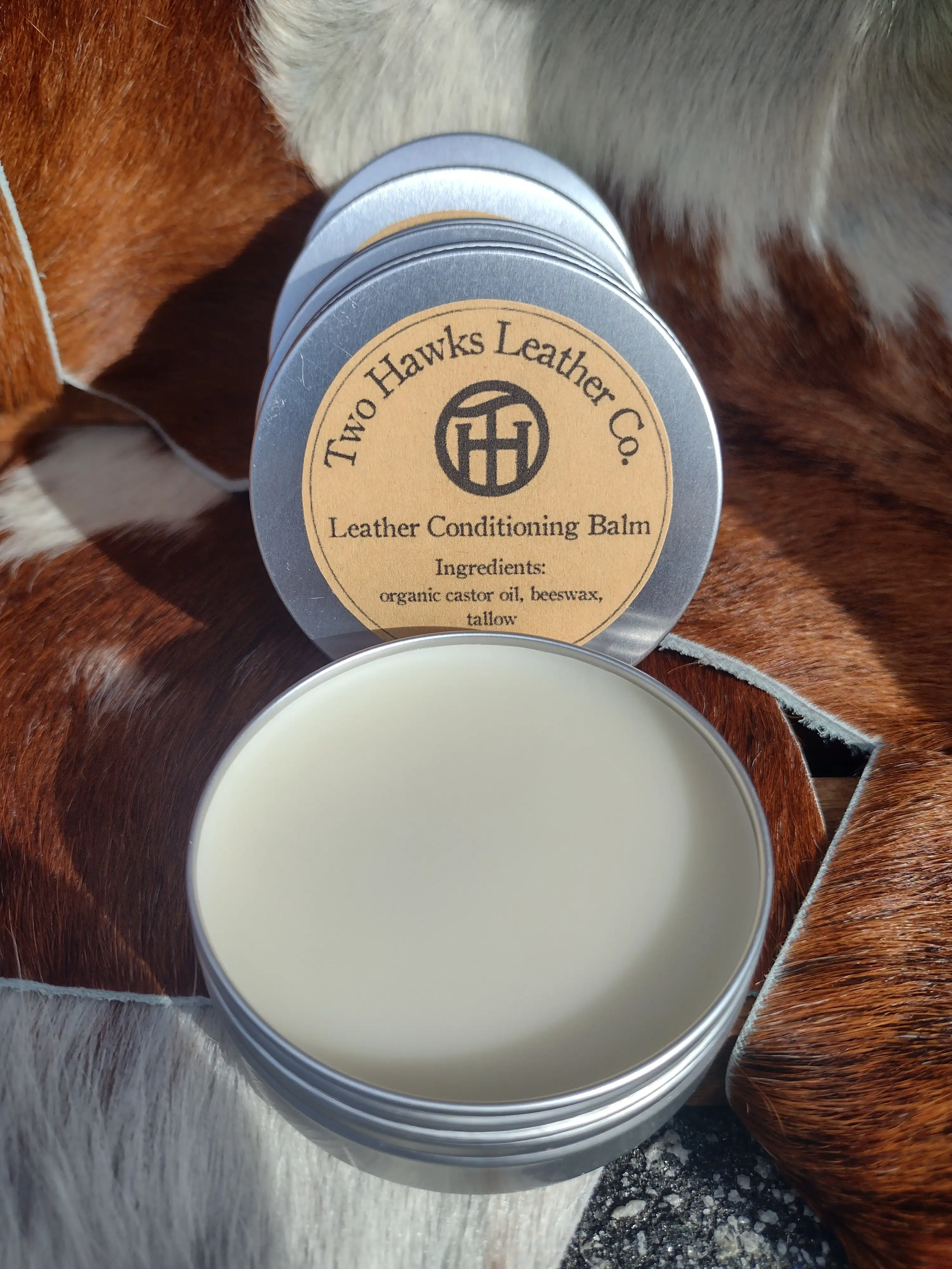 An open 2-ounce tin of leather balm revealing a smooth white balm inside, displayed alongside closed tins displayed on brown and white pelts.