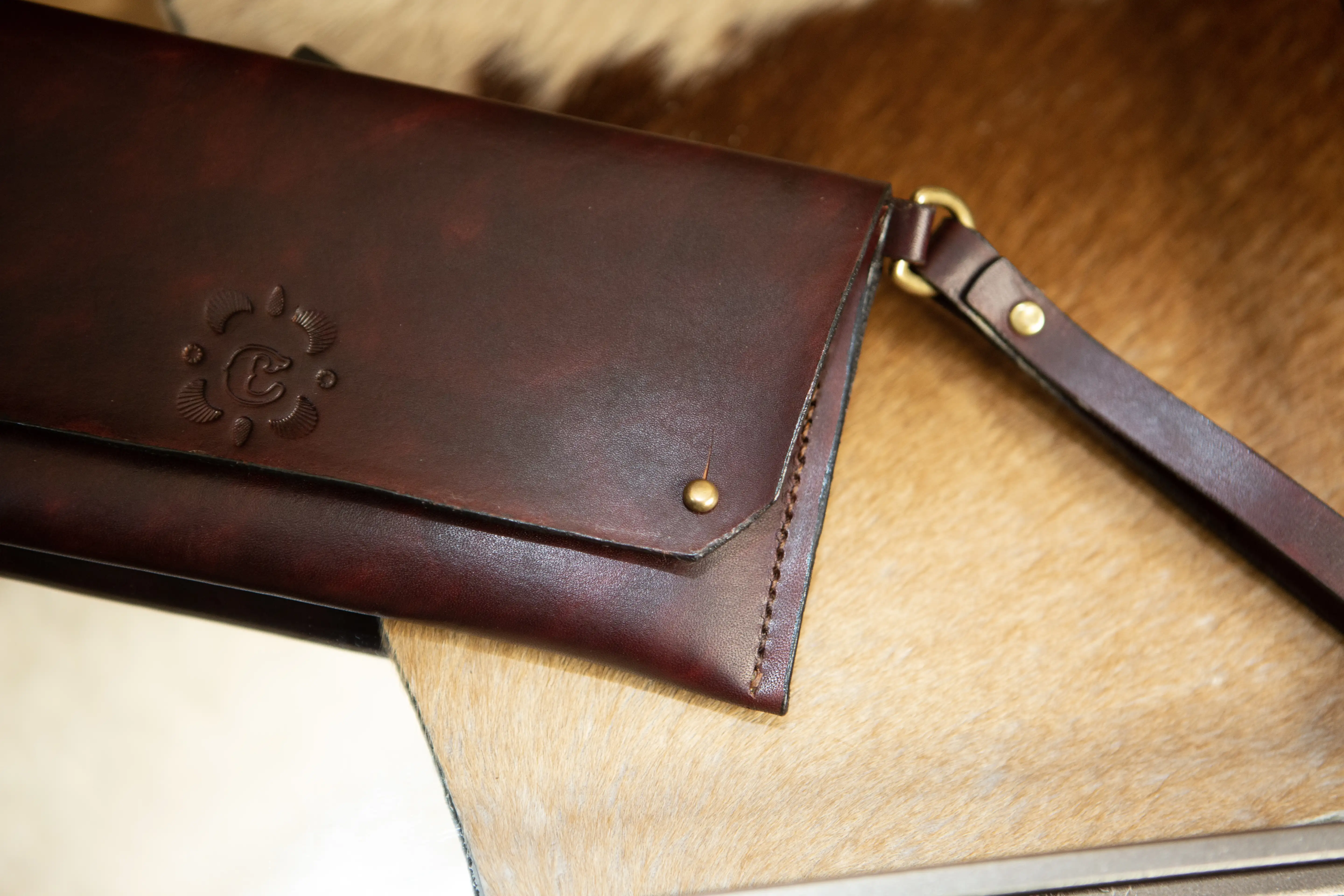 A dark brown leather clutch with a hand-stamped monogram on the front face displayed on a mirrored surface and a brown and white pelt.