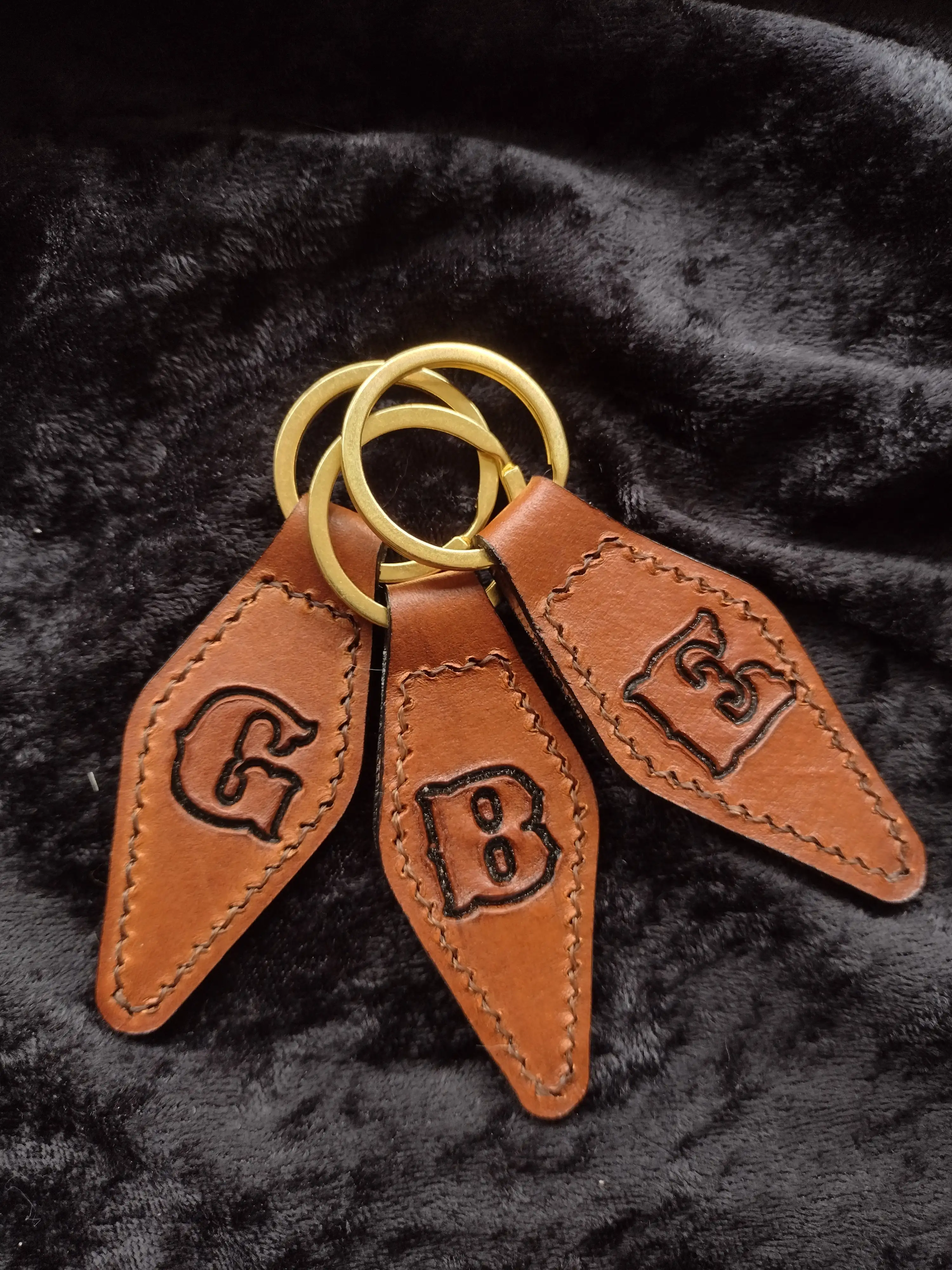 Three monogrammed leather keychains with bronze rings displayed on a dark fabric.