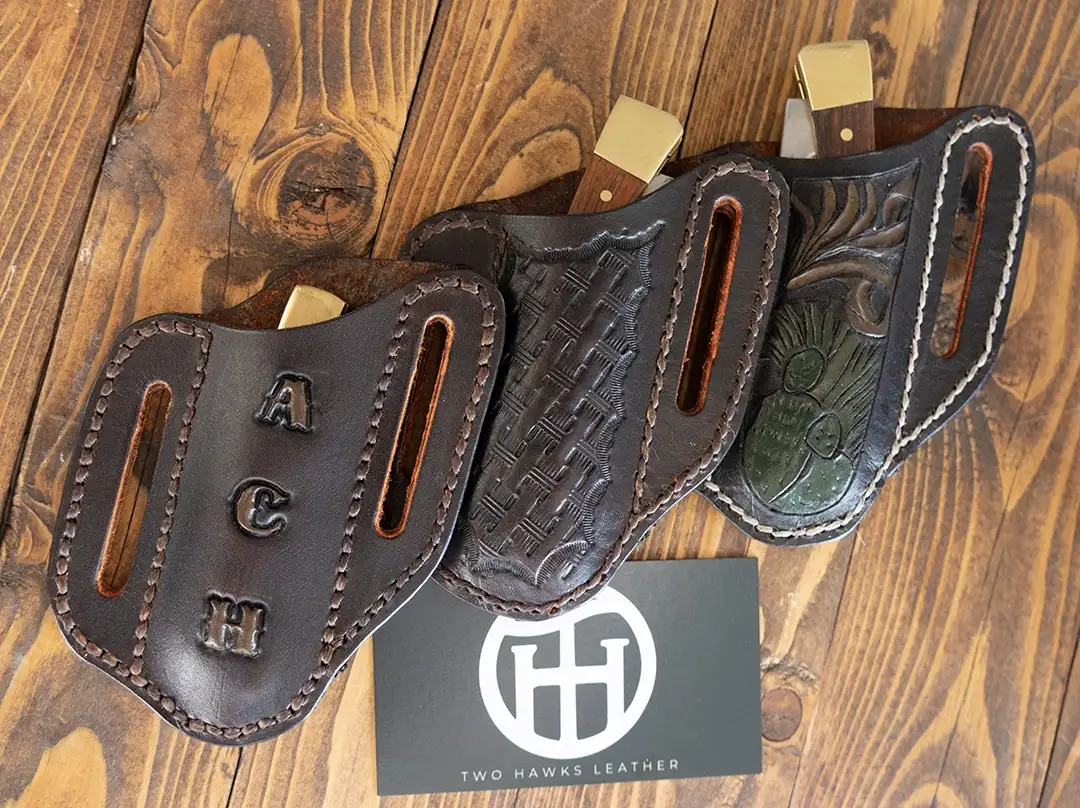 Three dark brown leather pancake sheaths displayed on a wooden panel — a monogrammed sheath on the left, a basketweave sheath in the center, and a hand-tooled nature scene sheath on the right.