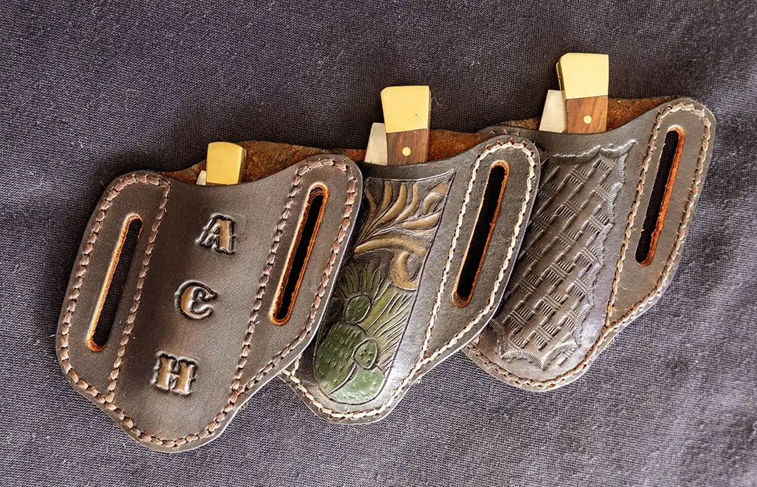 Three dark brown leather pancake sheaths displayed on black fabric — a monogrammed sheath on the left, a hand-tooled nature scene in the center, and a basketweave sheath on the right.