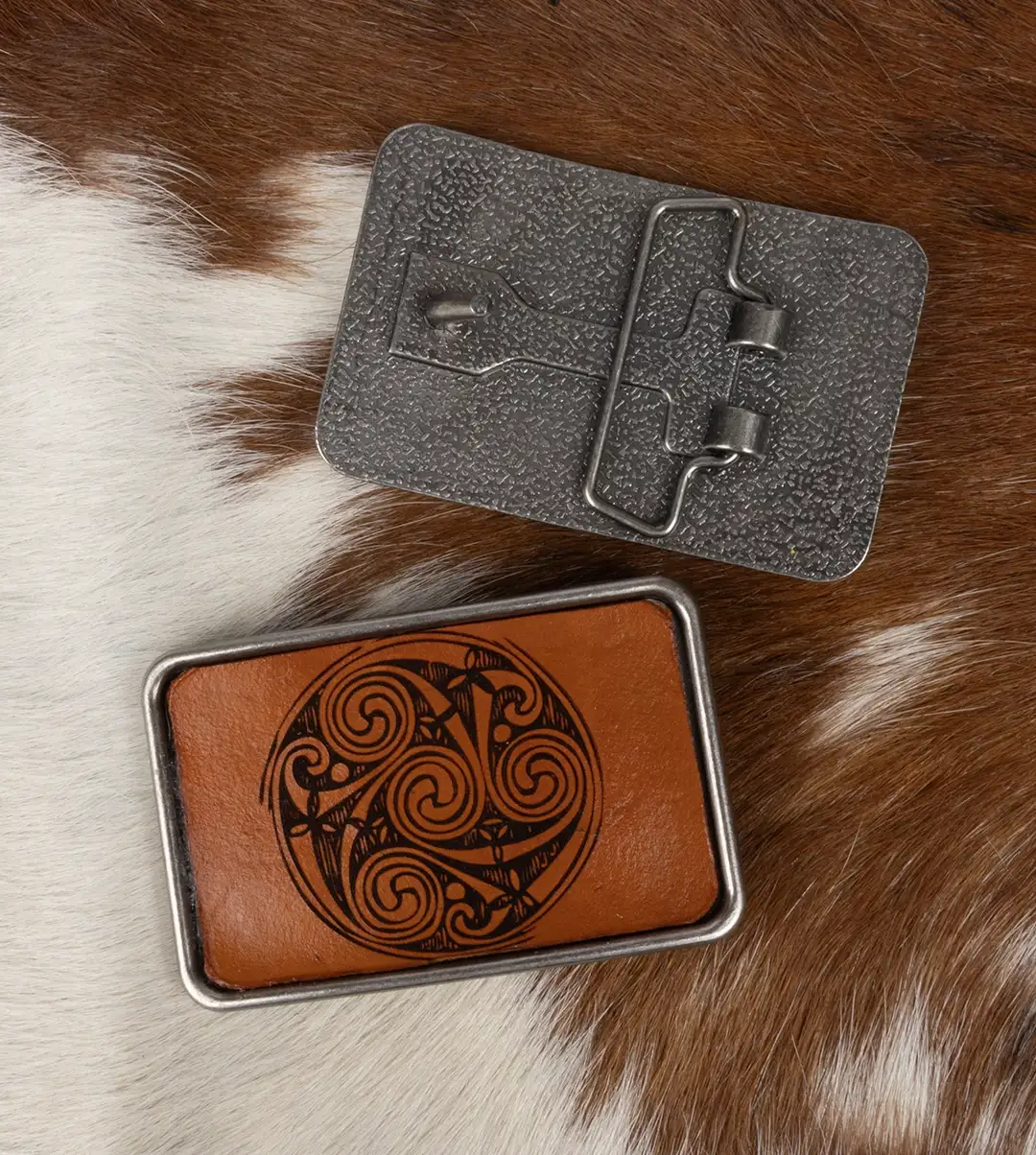 A silver belt buckle featuring a laser engraved leather inlay of a Pictish circle displayed on a brown and white pelt.