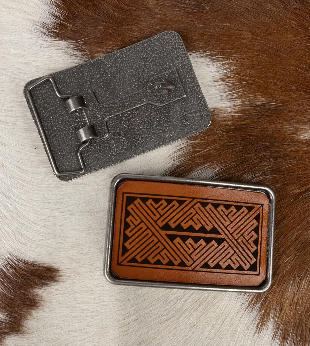 A silver belt buckle featuring a laser engraved leather inlay of Pictish knotwork displayed on a brown and white pelt.