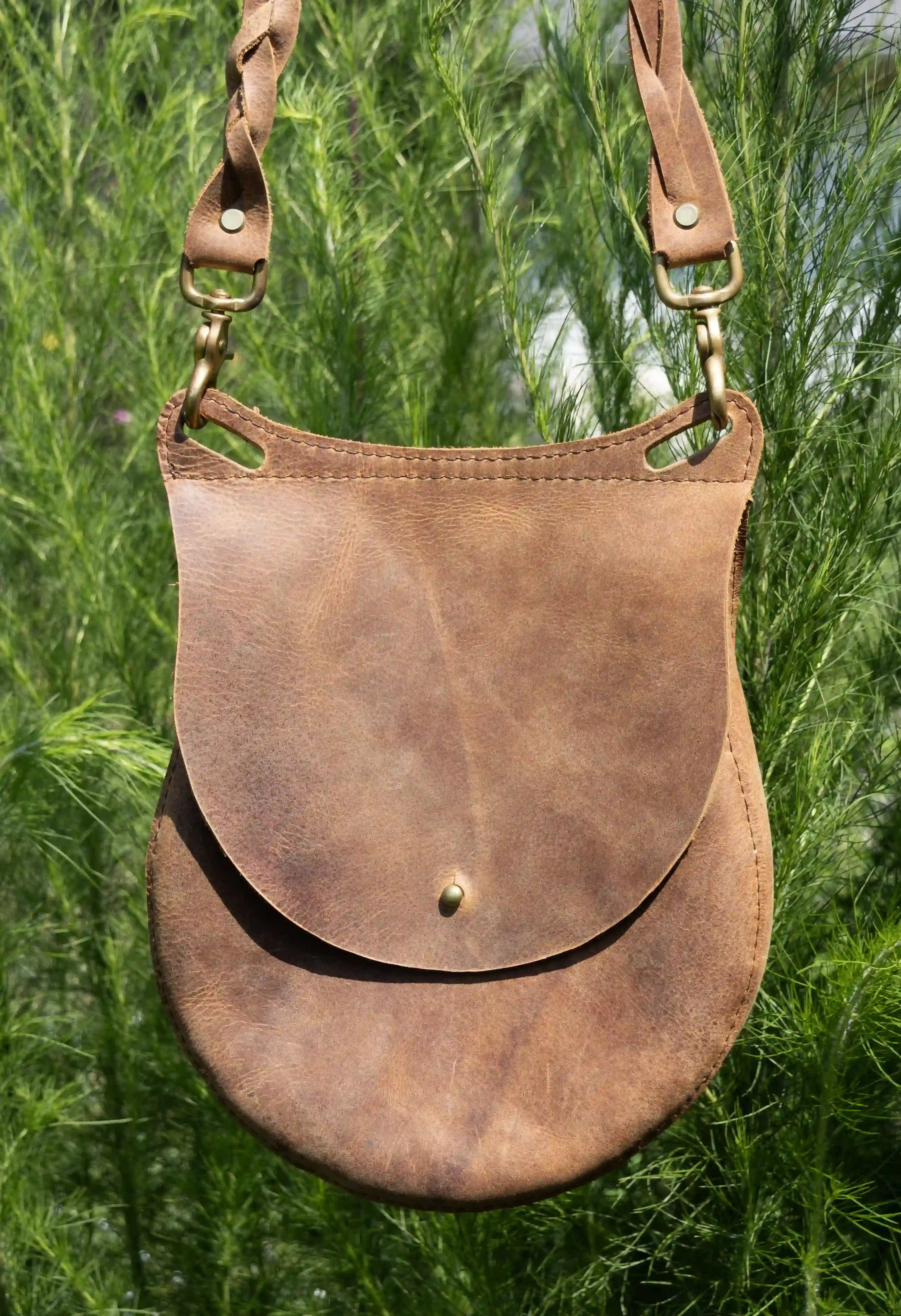 A tan leather shoulder bag with a removable woven strap and bronze hardware being held outdoors in front of trees.