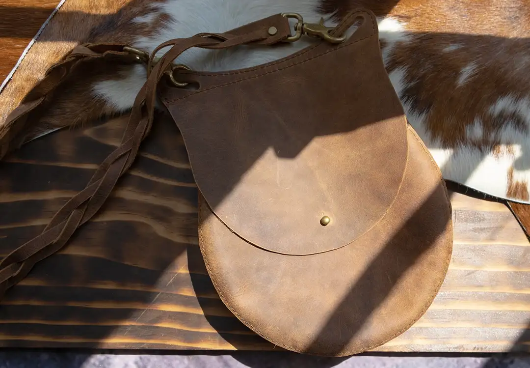 A tan leather shoulder bag with a removable woven strap and bronze hardware laying flat on a wooden table on top of a pelt.