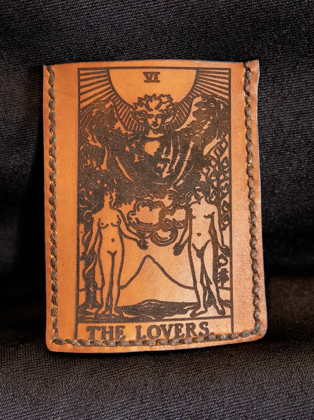 A tan leather card wallet featuring a hand-embossed image of The Lovers tarot card on the front face displayed on dark fabric.