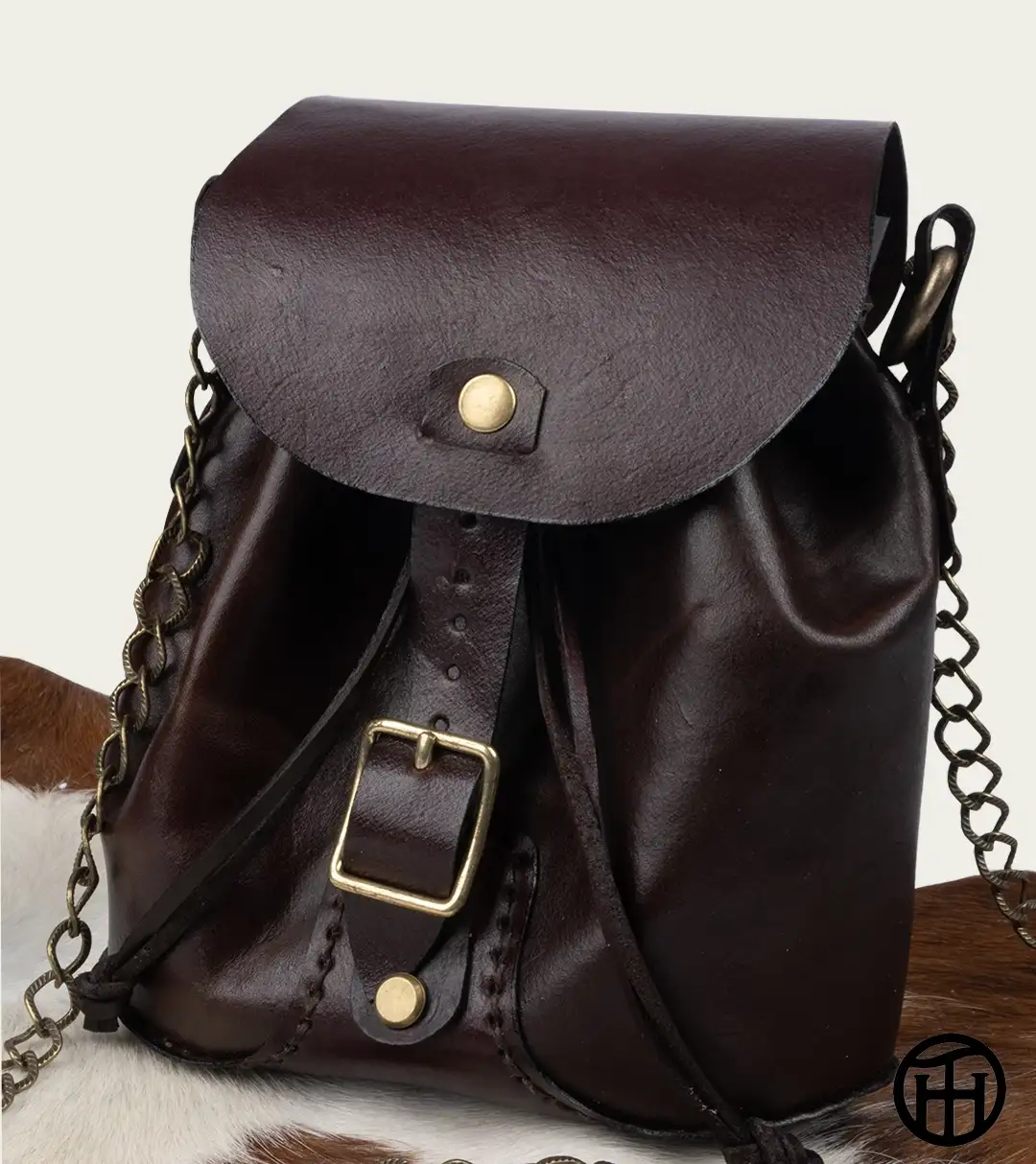 A dark brown leather cinch bag with a bronze buckle strap and metal chain shoulder strap displayed on a brown and white pelt against a neutral background.