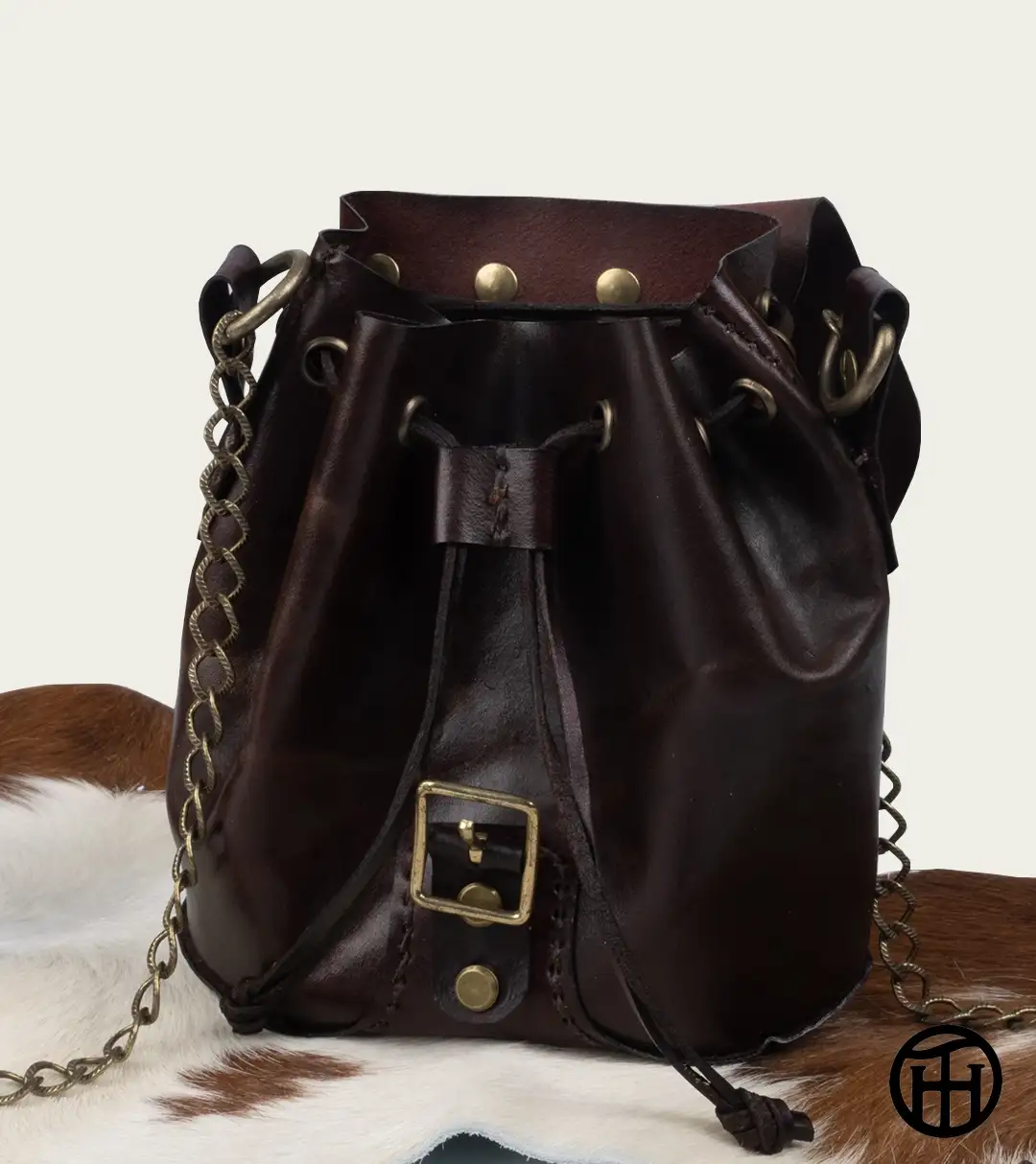 An open dark brown leather cinch bag, with the bronze buckle strap and metal chain shoulder strap visible displayed on a brown and white pelt against a neutral background.