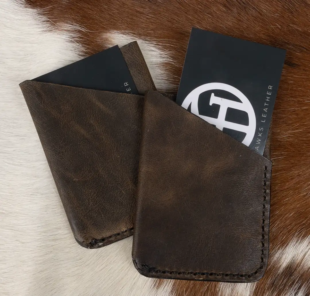 Two dark brown crazy horse leather three-pocket wallets with business cards in their pickets displayed on a brown and white pelt.