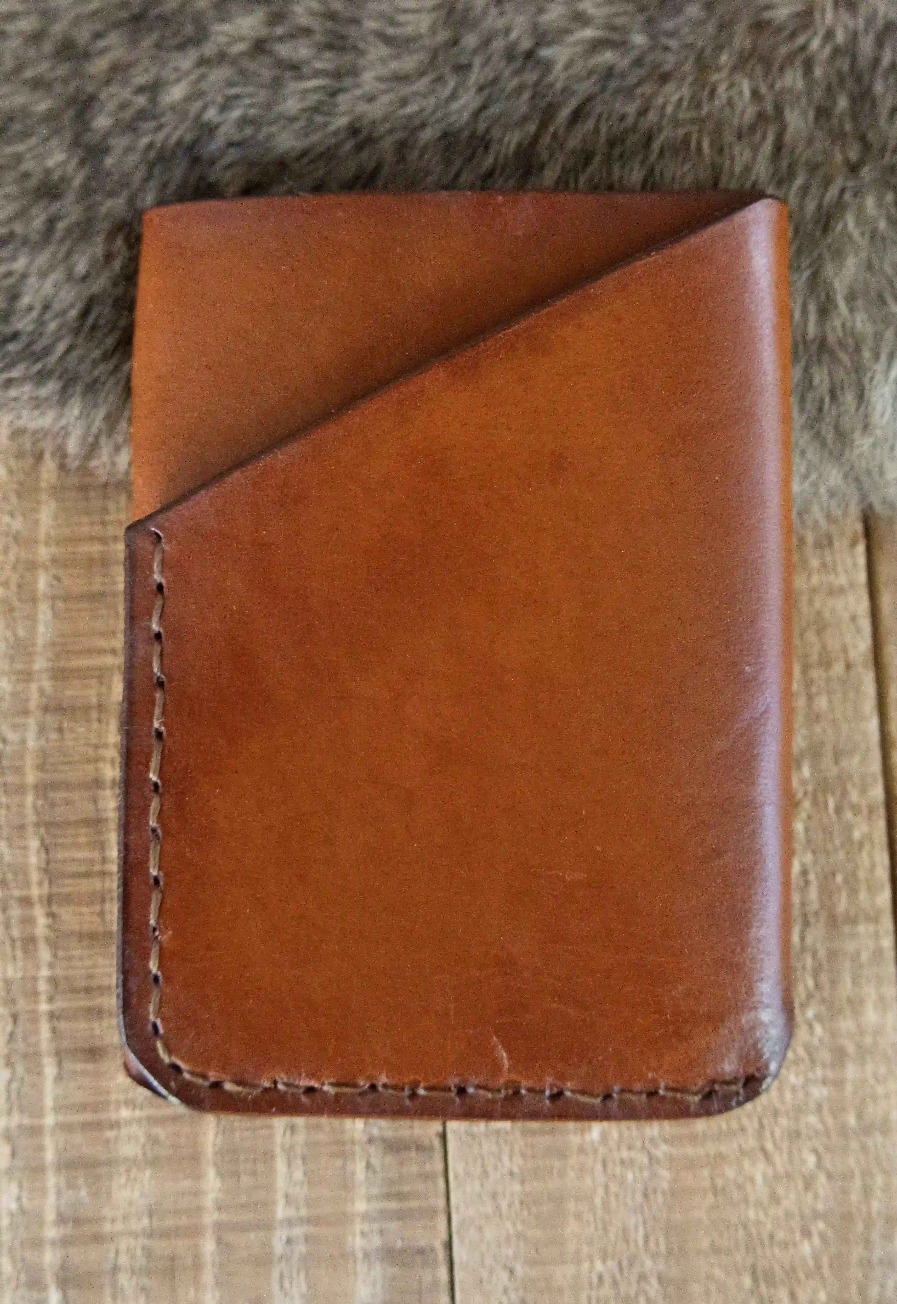 A light brown leather three-pocket wallet displayed on a wooden surface with a fur pelt.