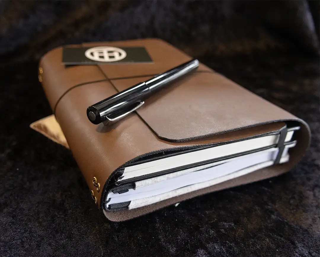A light brown leather traveler's journal with a black closure band and bronze hardware, resting on a black cloth with a black pen and business card placed on top.