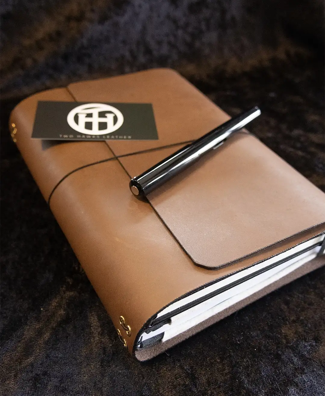 A light brown leather traveler's journal with a black closure band and bronze hardware, resting on a black cloth with a black pen and business card placed on top.
