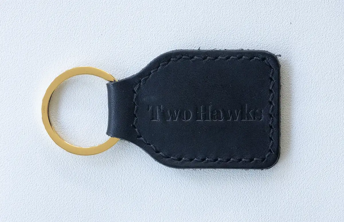 A black leather keychain stamped with the Two Hawks Leather logo and fitted with a bronze key ring displayed against a neutral background.